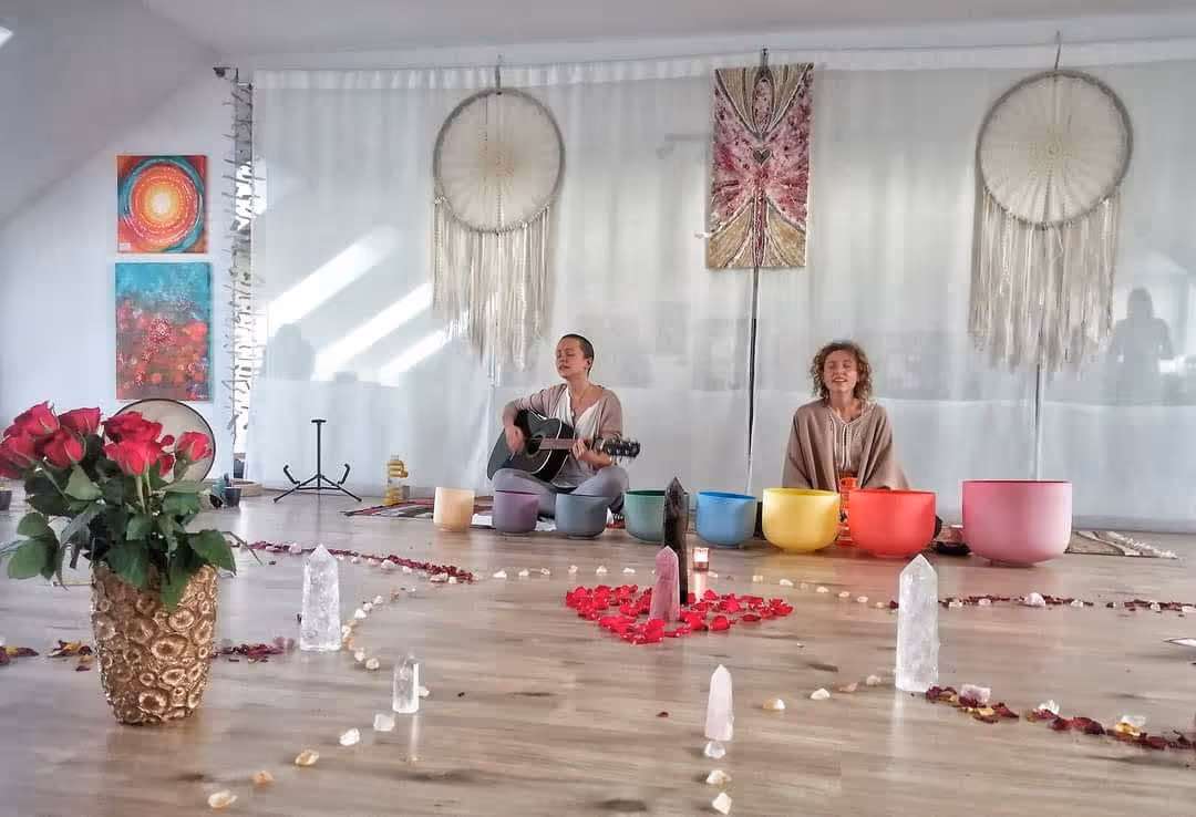 Rad farebných kryštálových spievajúcich mís, kryštálov a sviečok s lupeňmi ruží rozložený na drevenej podlahe; v pozadí sedia dvaja ľudia, jeden s gitarou.